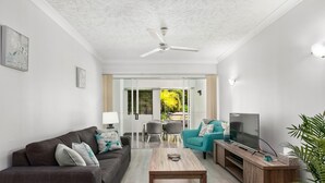 Een moderne woonkamer met een bank, salontafel en tv-meubel. Er is een plafondventilator, een eethoek met stoelen en uitzicht op groen buiten.