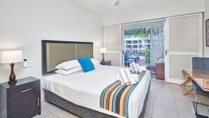 A hotel room with a bed, a desk, a chair, a ceiling fan, and a view of a building through the window. - Whispering Palm 2 Bedroom Beach Club Apartment (Palm Cove)