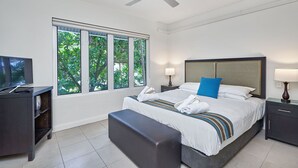 A hotel room with a bed, a TV, a window with a view of trees, a ceiling fan, and a nightstand with a lamp.
