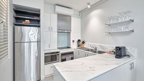 A modern kitchen with a stainless steel refrigerator, a microwave, an oven, and a coffee machine. There are white cabinets, a marble countertop, and a shelf with glasses.