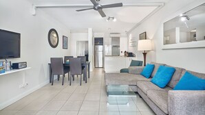A modern living room with a flat-screen TV, dining area, kitchen, and a ceiling fan.