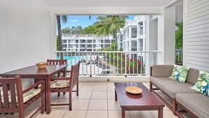 A balcony with a dining table, chairs, and a sofa. There is a pool area visible in the background.
