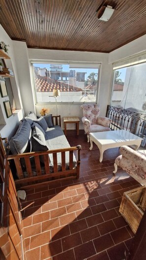 A sunlit room with a wooden ceiling, a sofa, two chairs, a small table, and a view of buildings through a large window. - Siirli Pansiyon (Seferihisar)