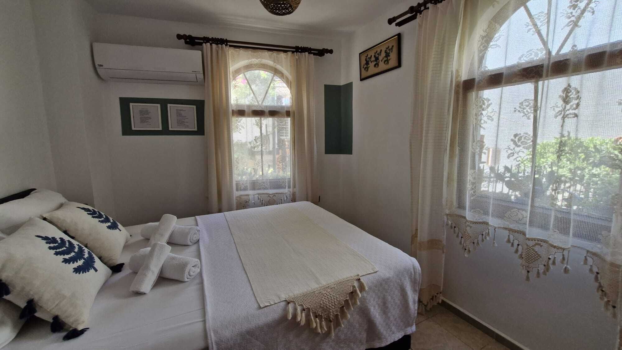 A neatly made bed with a white bedspread and a decorative fringe, a window with lace curtains, and an air conditioning unit on the wall.