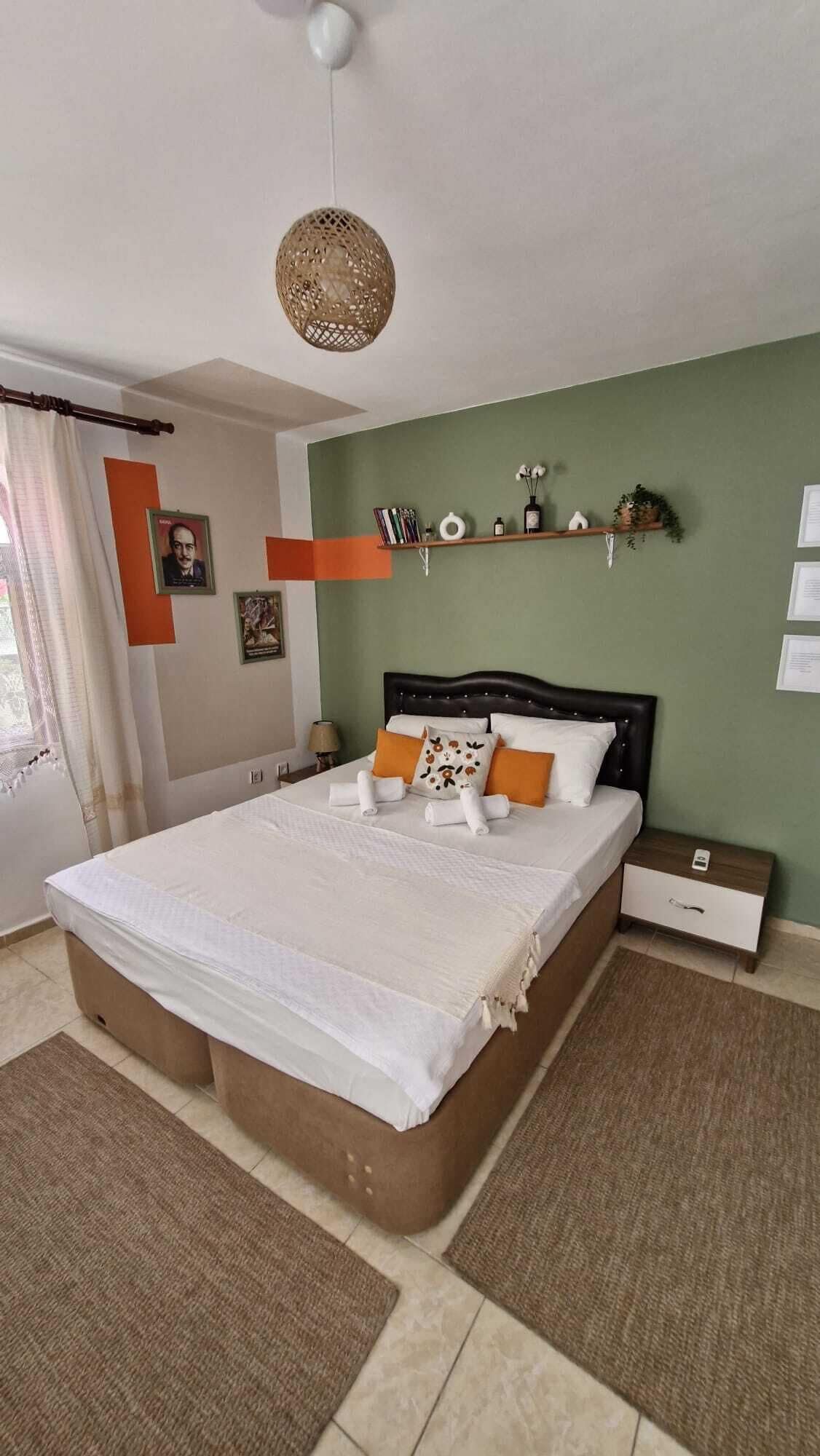 A bedroom with a large bed, a bedside table, a nightstand, a hanging light fixture, and a shelf with decorative items.