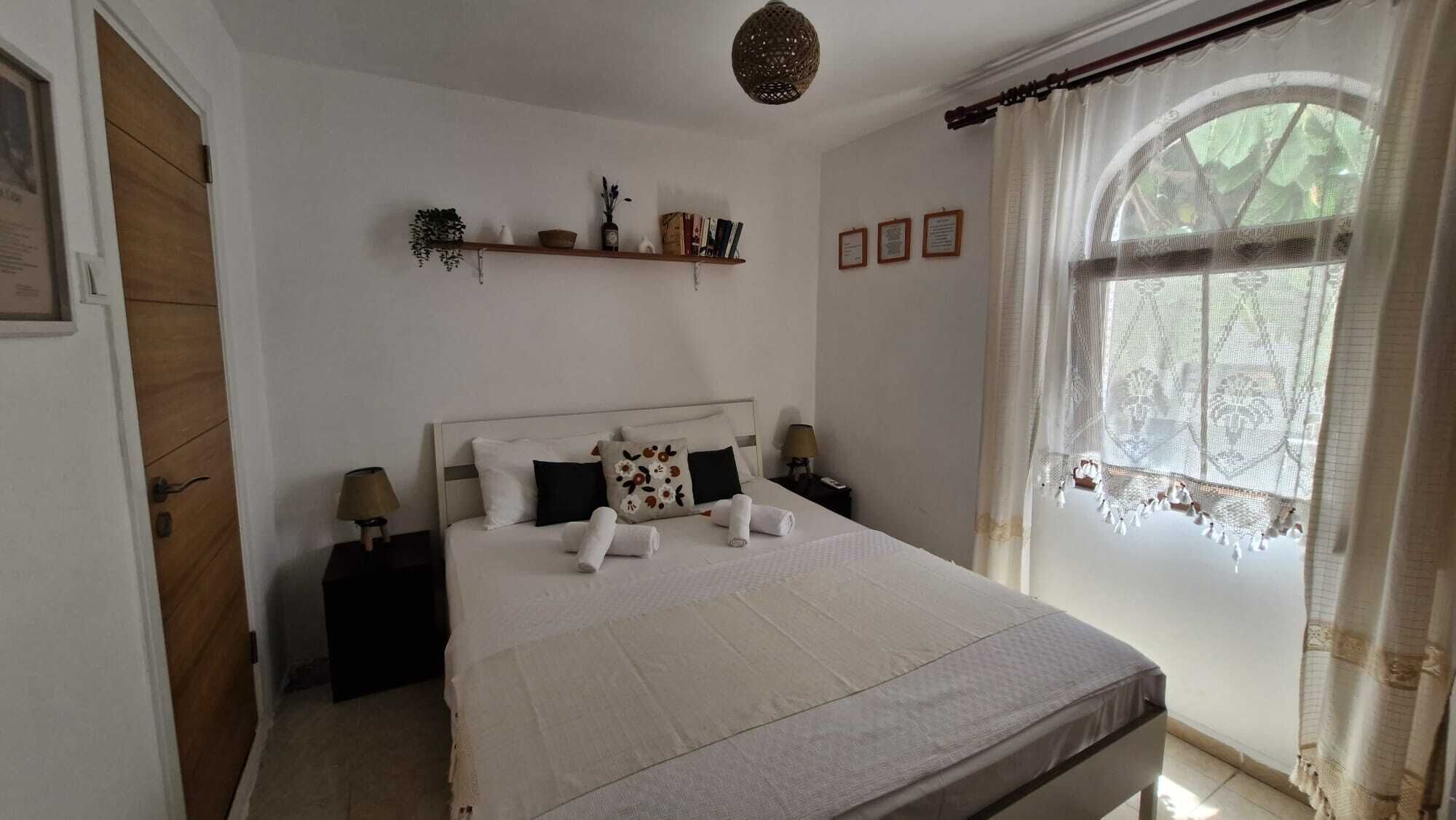 Une chambre à coucher bien rangée, avec un lit, des tables de chevet, une fenêtre avec des rideaux et une étagère contenant des livres et des objets décoratifs.