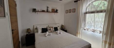 Une chambre à coucher bien rangée, avec un lit, des tables de chevet, une fenêtre avec des rideaux et une étagère contenant des livres et des objets décoratifs.