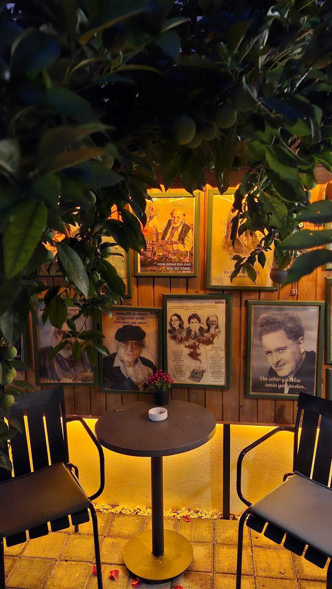 A cozy outdoor seating area with a round table and two chairs, surrounded by framed pictures and greenery.