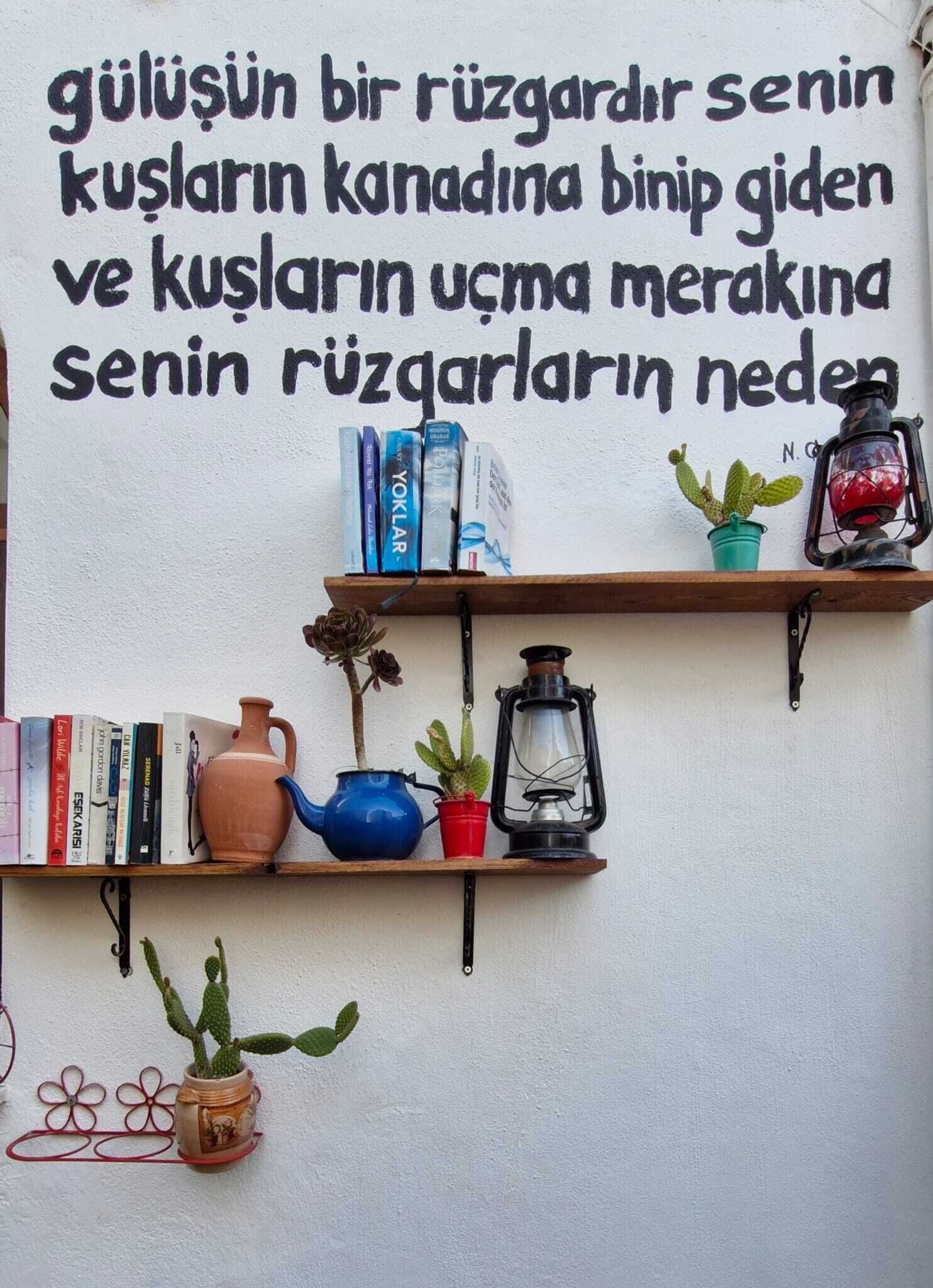 A wall with a Turkish quote and shelves with books, a teapot, a lantern, and potted plants.