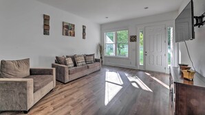 Een ruime woonkamer met een grijze bank, een kleinere fauteuil en een houten kast. Drie kunstwerken in lijsten hangen aan de muur, en een groot raam zorgt voor veel daglicht.