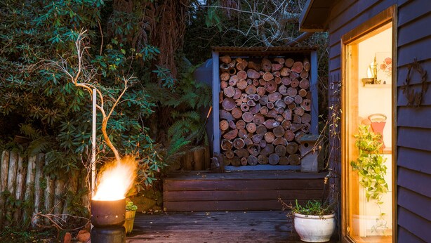 Een houten terras leidt naar een huis met een stapel houtblokken en een glazen deur waar een plantje in staat.