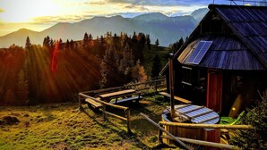 Eine Holzhütte mit geneigtem Dach und Solaranlage, umgeben von einem Holzzaun und einer Holzterrasse, mit Blick auf eine Berglandschaft im Abendrot.