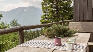Ein Holztisch mit kariertem Tischläufer, zwei weiße Tassen mit grünem Rand und eine rosafarbene Tasse mit Henkel, vor dem Hintergrund einer Berglandschaft und einer Kiefer.