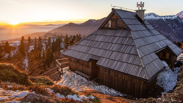 Eine Holzhütte mit geneigtem Dach, umgeben von schneebedeckten Bergen und Bäumen, im Abendlicht.