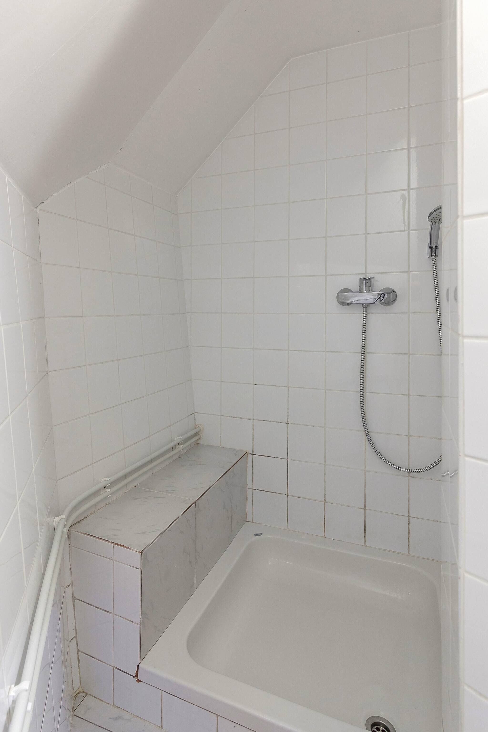 A small, white-tiled bathroom with a corner shower, a bathtub, and a visible drain.