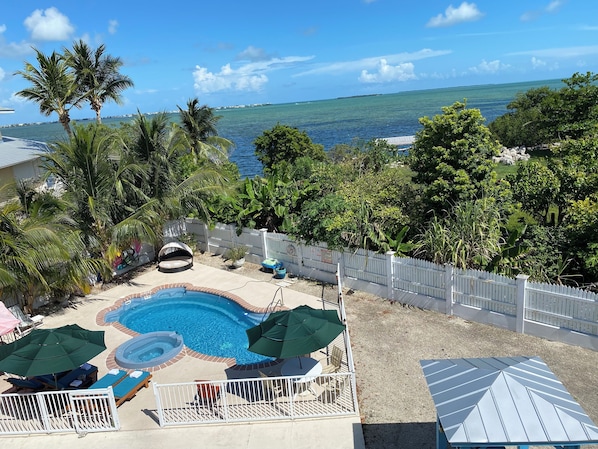 A swimming pool with lounge chairs and umbrellas, surrounded by a white fence and tropical vegetation. - Luxury Oceanview Pool Eco-friendly Villa Near Key West (Summerland Key)