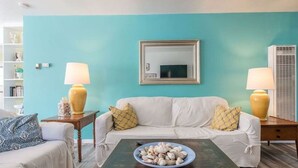 Un salon avec un mur de couleur turquoise, un canapé blanc, deux tables d’appoint avec des lampes, une table basse sur laquelle se trouve un saladier rempli de fruits, et un miroir au-dessus du canapé.