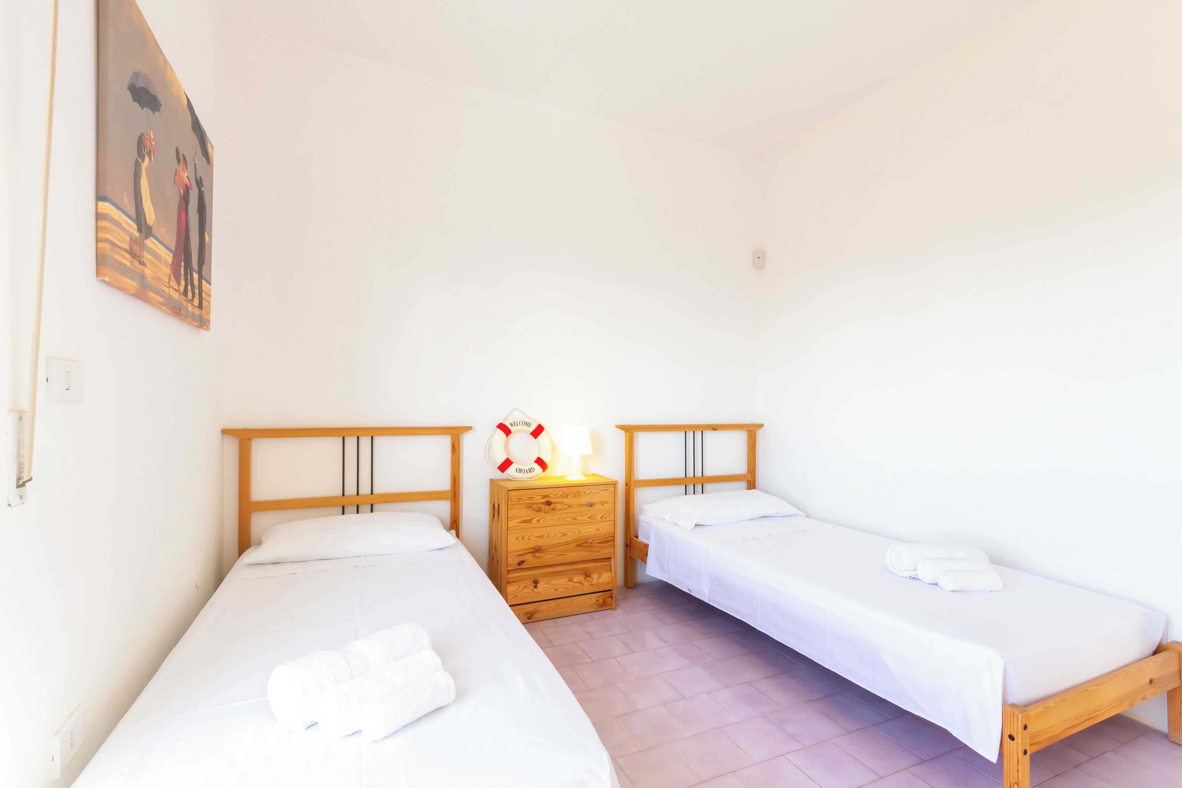 A room with two single beds, a wooden nightstand with a welcome sign, and a painting on the wall.
