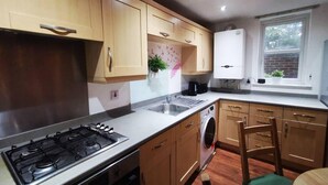 A modern kitchen with wooden cabinets, a gas stove, a washing machine, and a window with a view of greenery. - London - 2 Bed Flat - Sleeps 4 - Parking - Wifi (London)