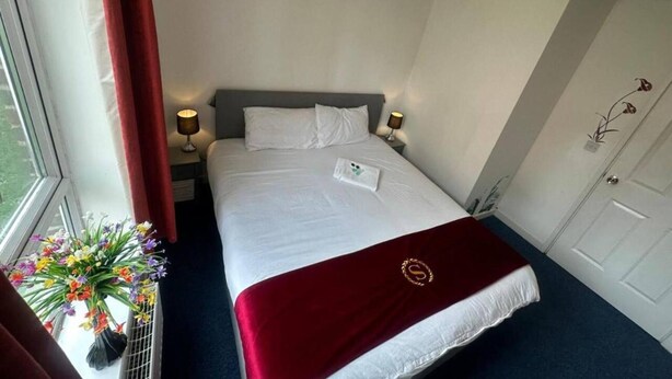 A hotel room with a bed, bedside lamps, a window with curtains, a door, and a small plant on the windowsill. - London - 2 Bed Flat - Sleeps 4 - Parking - Wifi (London)