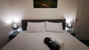 A neatly made bed with white linens, two pillows, and folded towels. There are two bedside lamps on either side of the bed. A painting of a landscape hangs above the bed. - London - 2 Bed Flat - Sleeps 4 - Parking - Wifi (London)