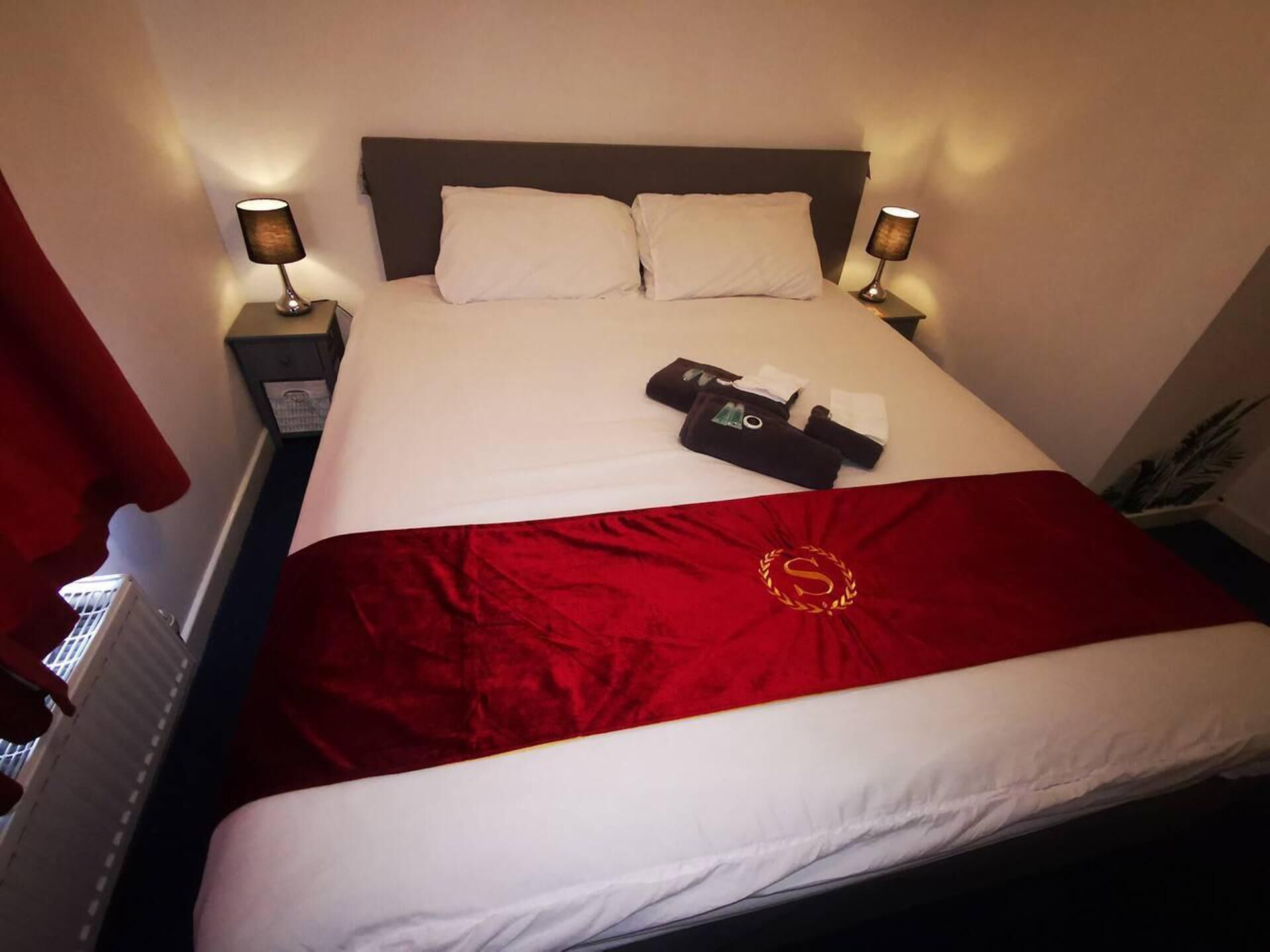 A hotel room with a neatly made bed, two bedside lamps, and a red throw blanket with a logo.