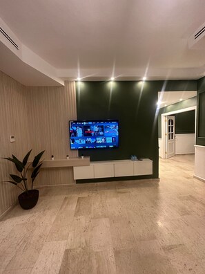 En moderne stue med et fladskærms-tv monteret på væggen, et hvidt tv-møbel nedenunder, en potteplante og en gang der fører til et andet rum.