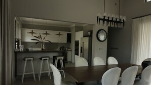 A modern dining area with a wooden table and white chairs, a kitchen in the background, and a wall clock. - Royal Park Villa De Luxe 3 Chambres (Balaclava)