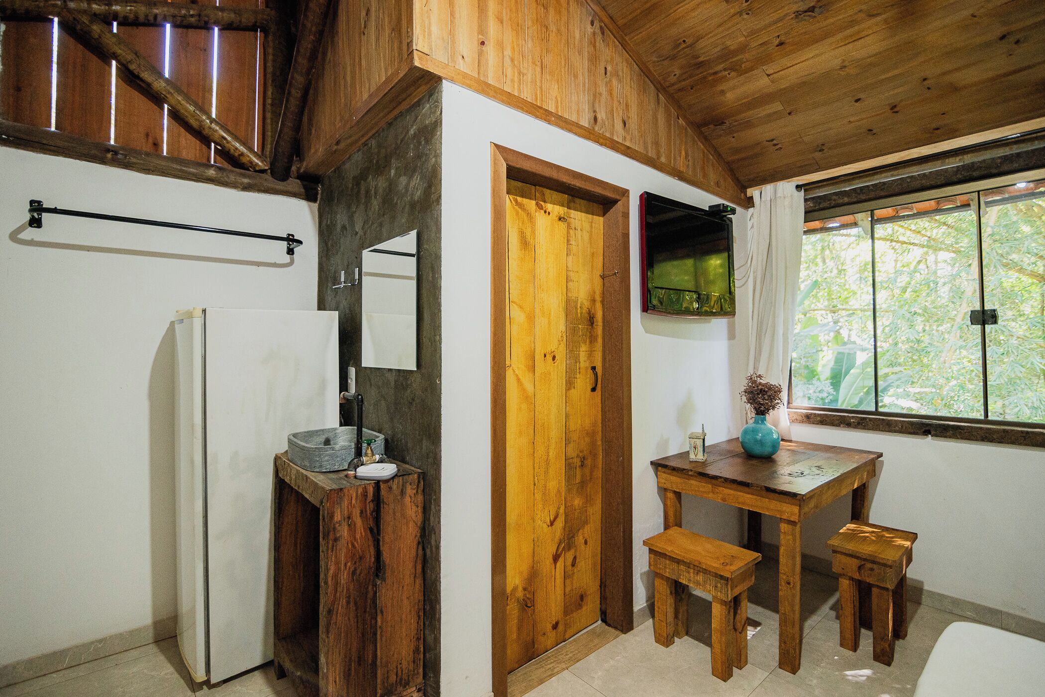 A rustic kitchen with wooden beams, a wooden door, a small table with a vase, and a window with a view of greenery.
