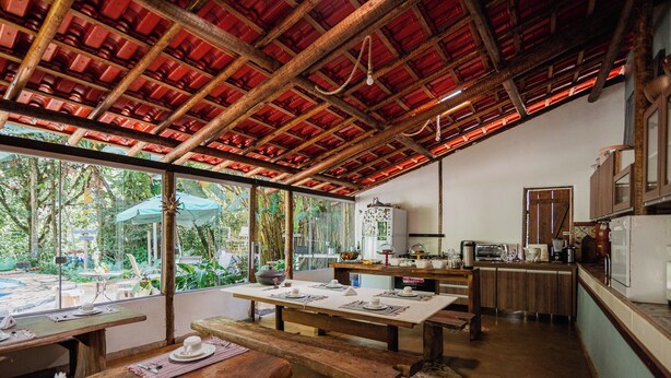 A rustic dining area with wooden tables and benches, a kitchen with basic appliances, and large windows offering a view of the outdoors. - Pousada Passaredo (Brumadinho)