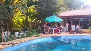A pool area with a tiled deck, surrounded by greenery and a building with a tiled roof. - Pousada Passaredo (Brumadinho)