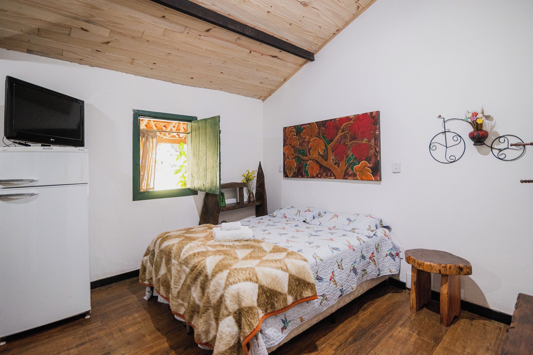Un dormitorio con cama, televisor sobre un mueble, ventana con cortina, un cuadro y una bicicleta decorativa colgada en la pared.