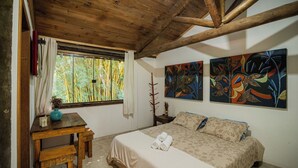 A bedroom with a wooden ceiling, a bed with a beige bedspread, two paintings on the wall, and a window with curtains. - Pousada Passaredo (Brumadinho)
