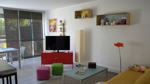 Ein modernes Wohnzimmer mit Flachbildfernseher, einem roten Schrank, einem grünen Hocker, einem gelben Sofa und einem Essbereich mit Glastisch und Stühlen.