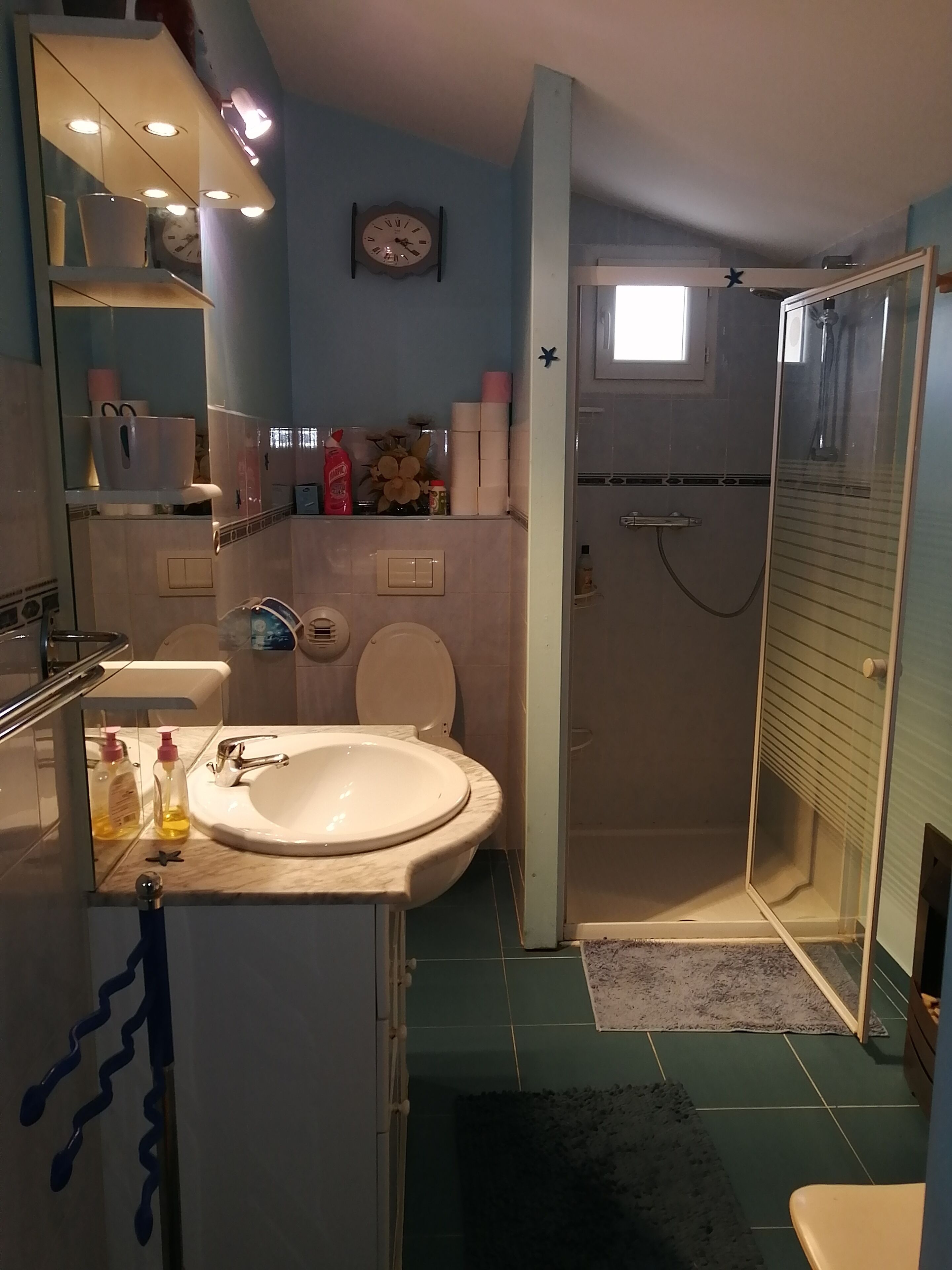 A bathroom with a sink, toilet, and shower. There is a clock on the wall and a mirror above the sink.