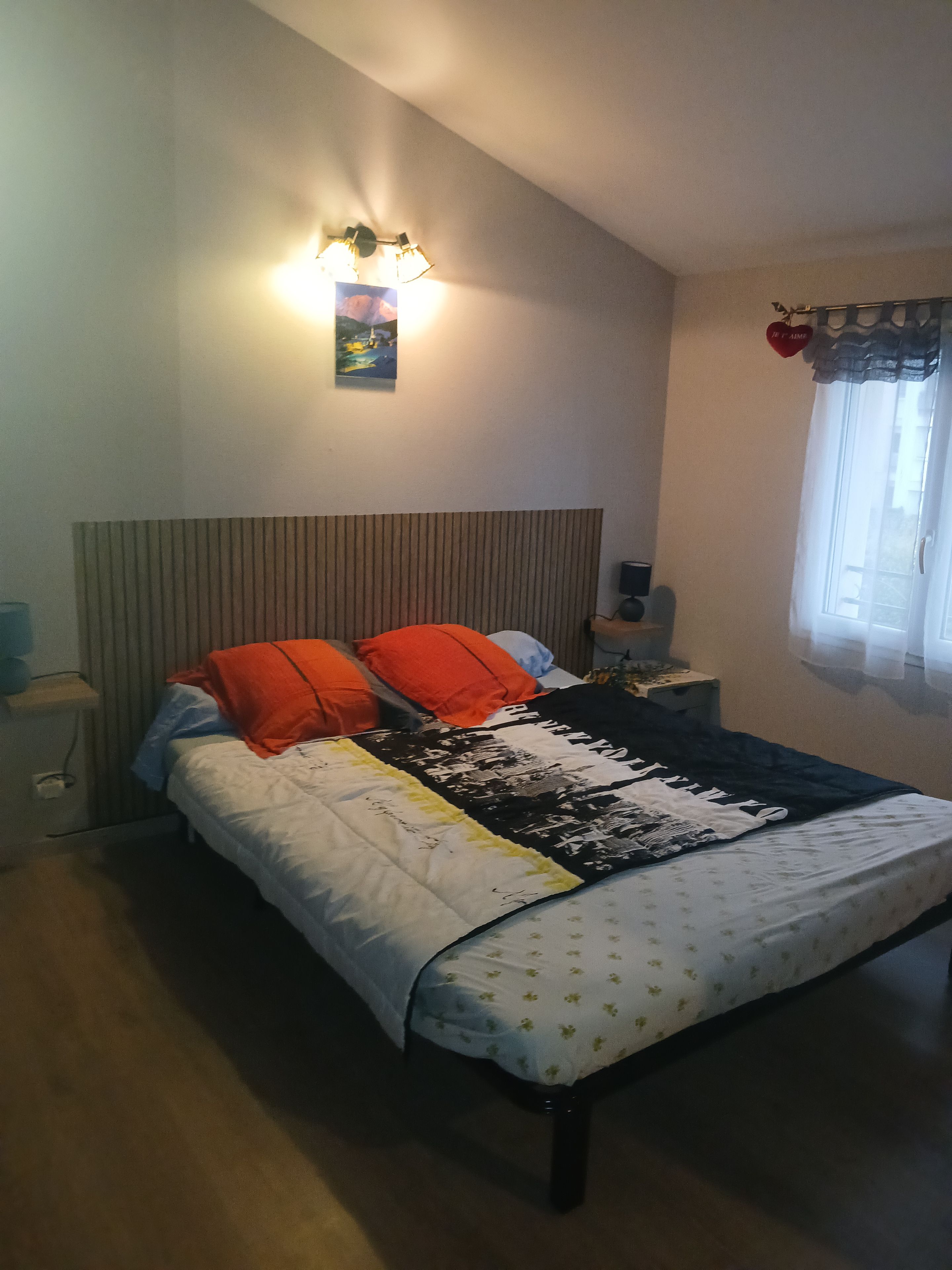 Un dormitorio con cama, mesita de noche, lámpara de pared y ventana con cortinas.
