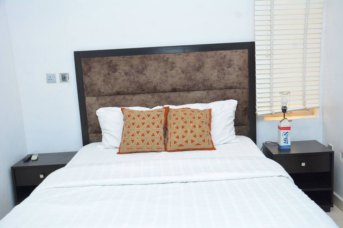 A neatly made bed with two decorative pillows, a bedside table with a lamp, and a power outlet on the wall.