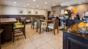 A hotel dining area with a long table, chairs, a cushioned bench, and a kitchen counter with appliances.