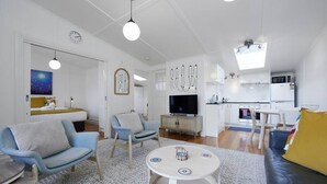 En moderne stue med sofa, lænestole, sofabord og tv. Der er et køkkenområde med mikrobølgeovn og spisebord.