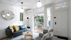 En moderne stue med en grå sofa, et rundt sofabord og en grå lænestol. Der er et stort spejl på væggen, en gulvlampe og et maleri. Stuen har store vinduer og en dør, der fører ud.