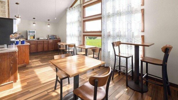 A restaurant with wooden tables and chairs, a kitchen area, and large windows with curtains. - Days Inn by Wyndham Wauseon (Wauseon)