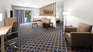 A hotel room with a blue patterned carpet, a sofa, a chair, a desk with a chair, a TV, a lamp, and a staircase. - Days Inn by Wyndham Wauseon (Wauseon)