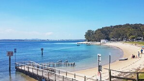 A beach with a wooden pier extending into calm waters, a sandy shore, and a forested area in the background. - Ocean Shores, 5 27 Weatherly Close - waterfront unit with views to Shoal Bay and wifi (Nelson Bay)