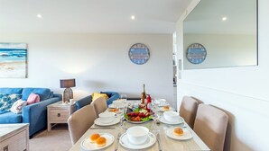 A dining area with a table set for four, a blue sofa, a lamp, and two wall clocks. - Ocean Shores, 5 27 Weatherly Close - waterfront unit with views to Shoal Bay and wifi (Nelson Bay)