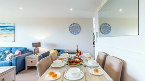 A dining area with a table set for four, a blue sofa, a lamp, and two wall clocks. - Ocean Shores, 5 27 Weatherly Close - waterfront unit with views to Shoal Bay and wifi (Nelson Bay)