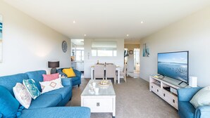 A modern living room with a blue sofa, a coffee table, and a TV. There's a dining area with a table and chairs, and a kitchen in the background. - Ocean Shores, 5 27 Weatherly Close - waterfront unit with views to Shoal Bay and wifi (Nelson Bay)