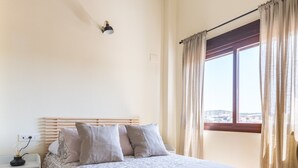 A bedroom with a wooden floor, a bed with a white and beige bedspread, a nightstand, a window with curtains, and a ceiling with exposed beams.