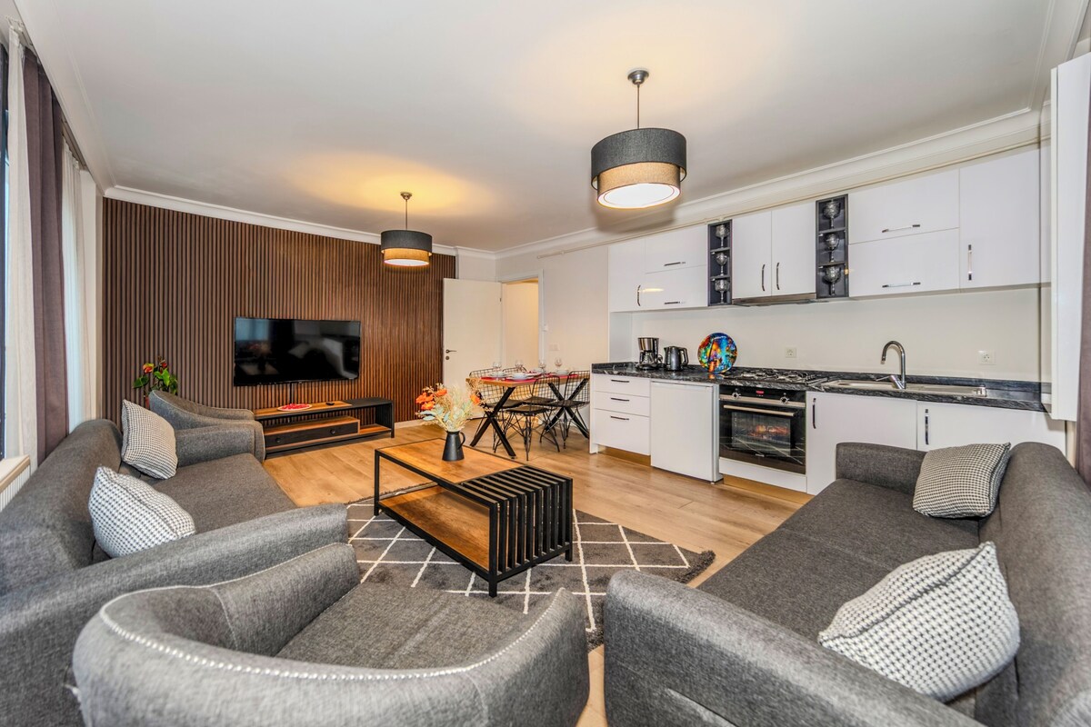 A modern living room with grey sofas, a coffee table, and a TV. There's a dining area with a table and chairs, and a kitchen with white cabinets and a stove.