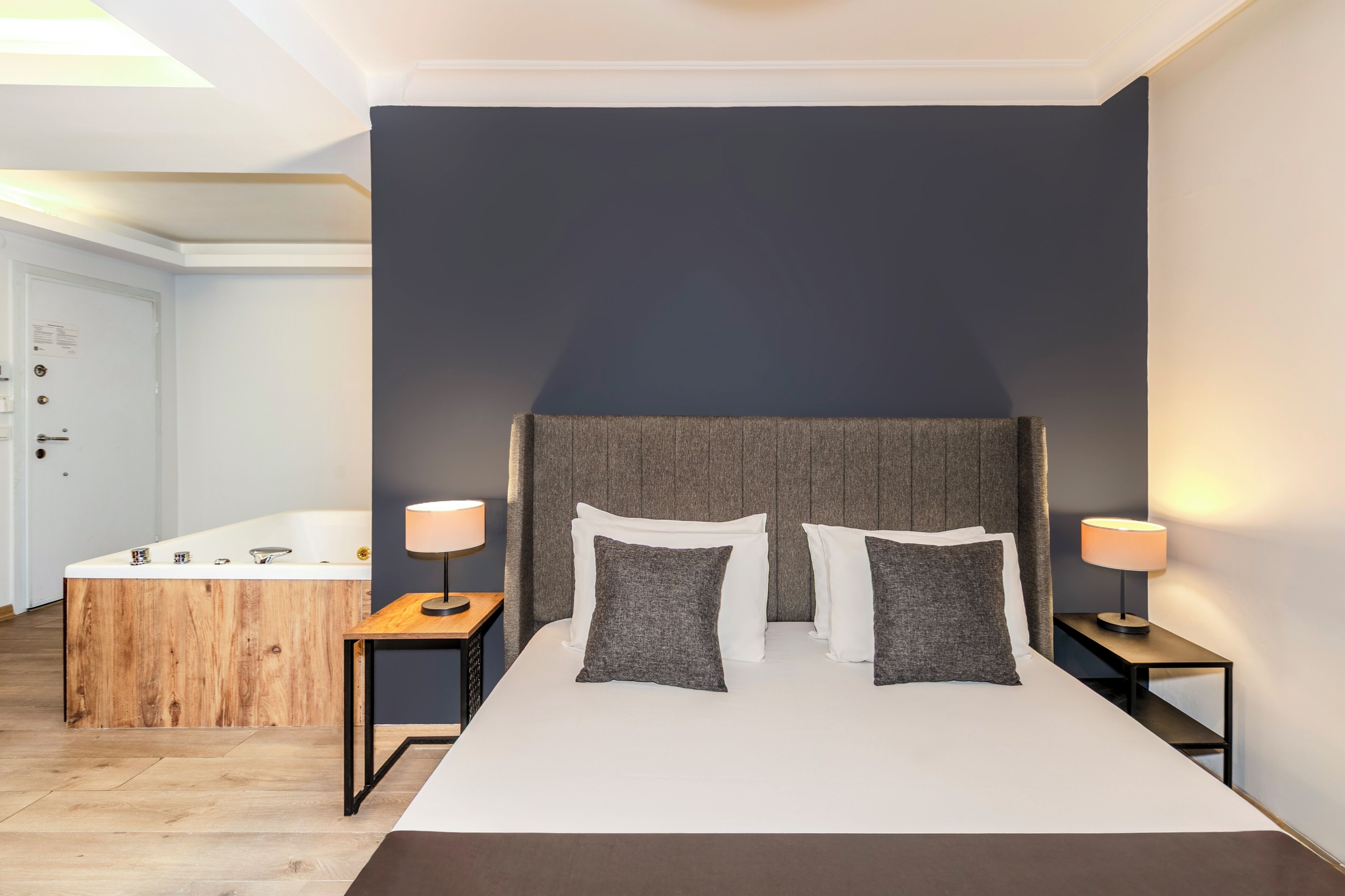 A modern hotel room with a large bed, two bedside tables with lamps, a wooden side table, a built-in bathtub, and a dark accent wall.