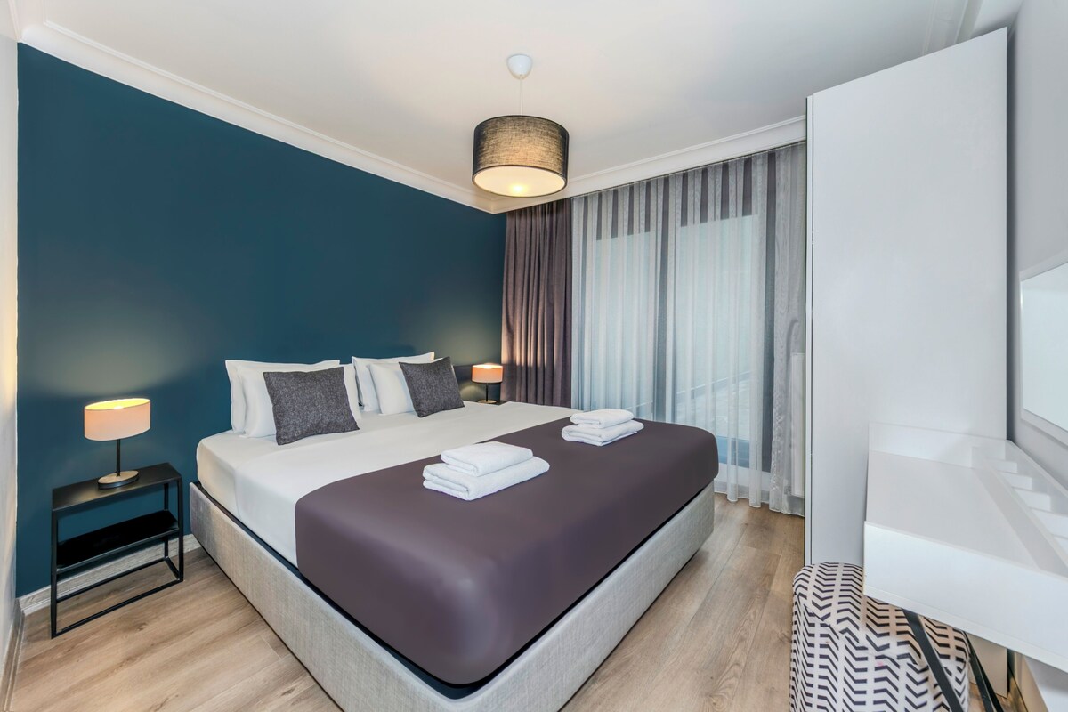 A modern hotel room with a large bed, a nightstand with a lamp, a TV, and a view of the outdoors.